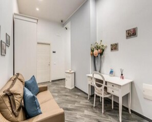 A modern hotel room with a wooden floor, a white desk, a white chair, a beige sofa with blue cushions, and a white door. - Agorà Borsa (Naples)