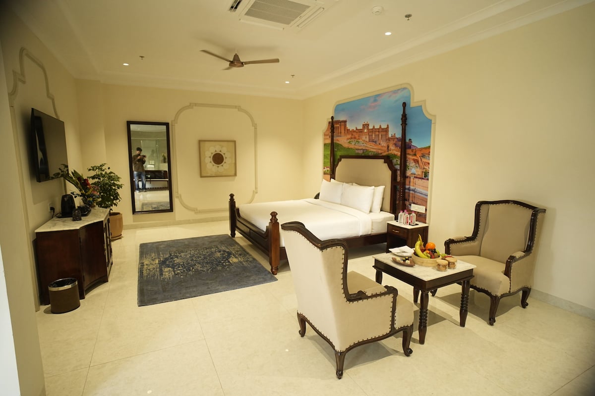 A hotel room with a bed, two armchairs, a small table with fruit, a mirror, a painting, and a ceiling fan.