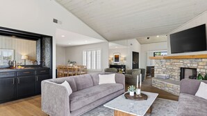 A spacious living area with a fireplace, a television mounted above it, and a kitchen area with dark cabinetry and a wooden ceiling.