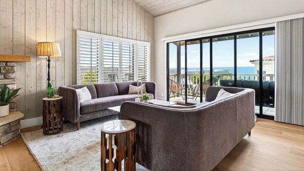 A modern living room with a grey sofa, a small side table, and a lamp. There is a large window with blinds, and a view of the sea is visible through the window.