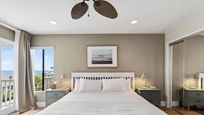 A bedroom with a large bed, two bedside tables with lamps, a ceiling fan, a framed picture on the wall, and a view through a window.