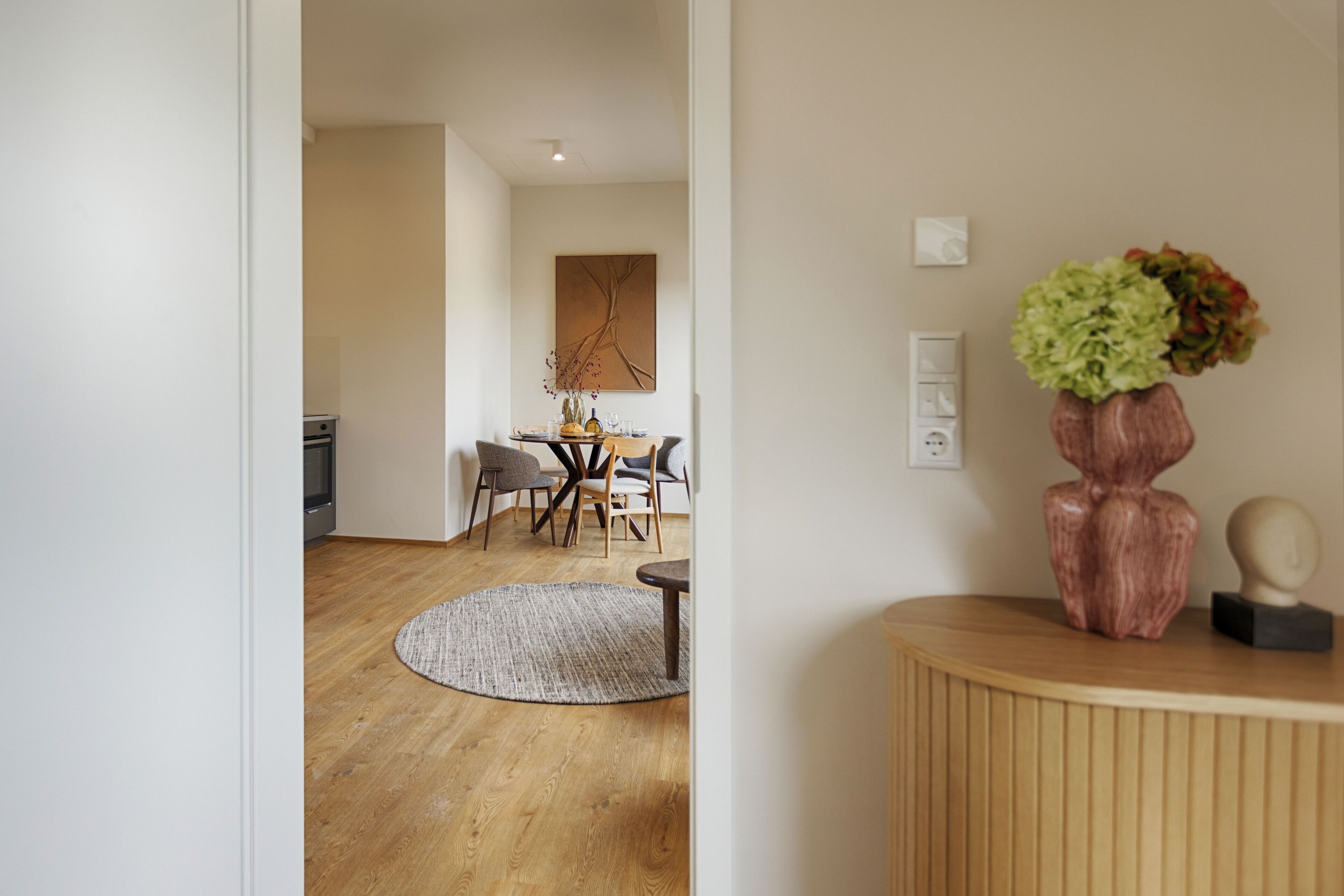 A modern living room with a wooden floor, a round rug, a dining table with chairs, and a vase with flowers on a wooden console table.