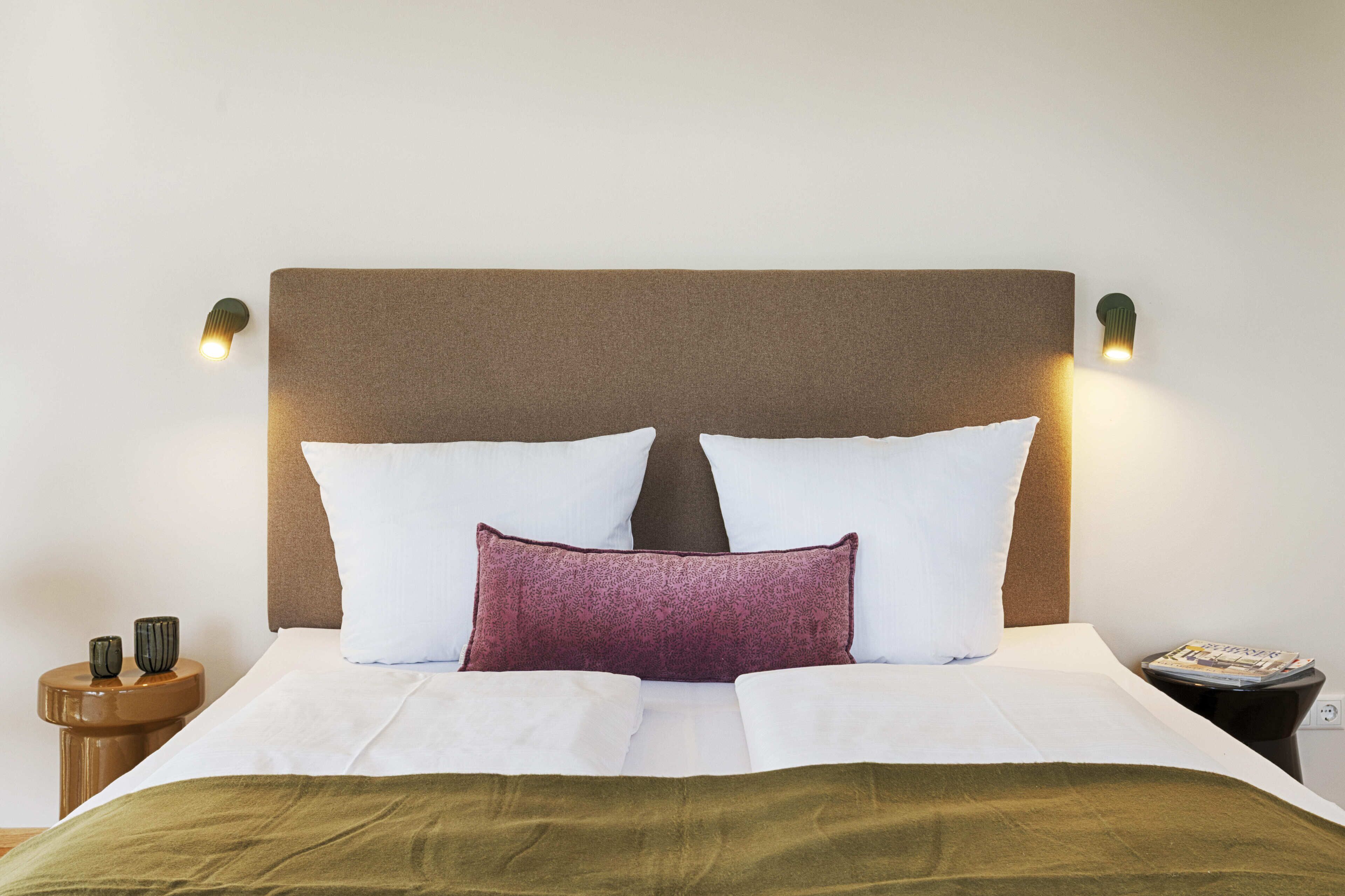 A neatly made bed with white and green bedding, a brown headboard, and two wall-mounted reading lamps.