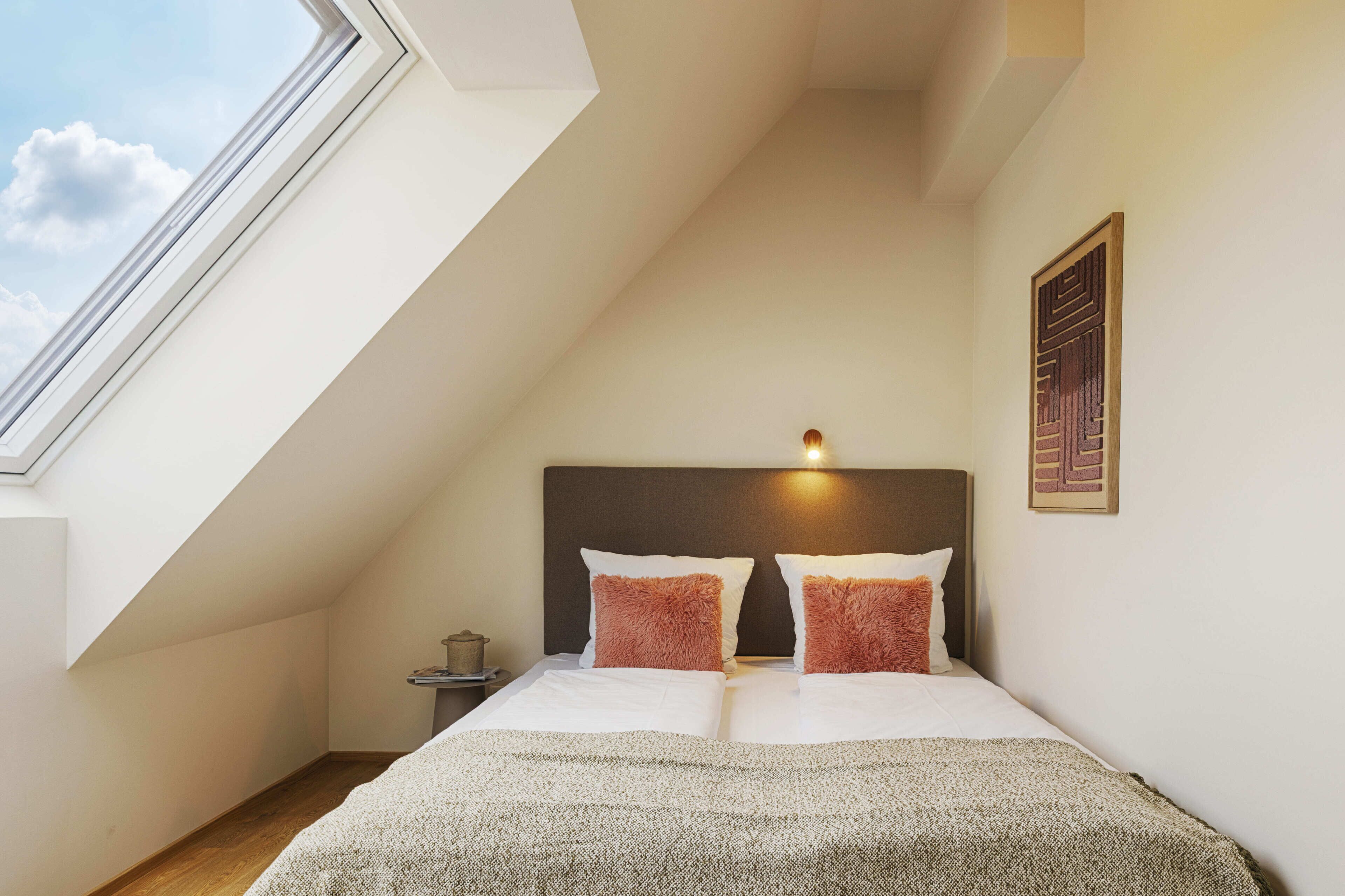 A bedroom with a sloped ceiling, a bed with white bedding and two decorative pillows, a bedside table with a lamp, and a framed artwork on the wall.