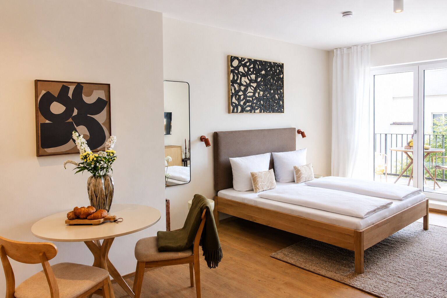 A modern hotel room with a bed, a round table with a vase and croissants, a chair, and a balcony with outdoor furniture.