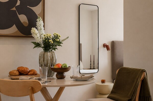 A dining area with a round table, a vase with flowers, a bowl of fruit, and a mirror on the wall.