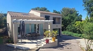Ein zweistöckiges Haus mit Terrasse, Sitzgelegenheiten im Freien und Blumentöpfen.