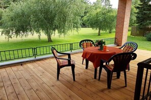 En træterrasse med et bord og stole, omgivet af gelænder i metal, med et træ og græs i baggrunden.