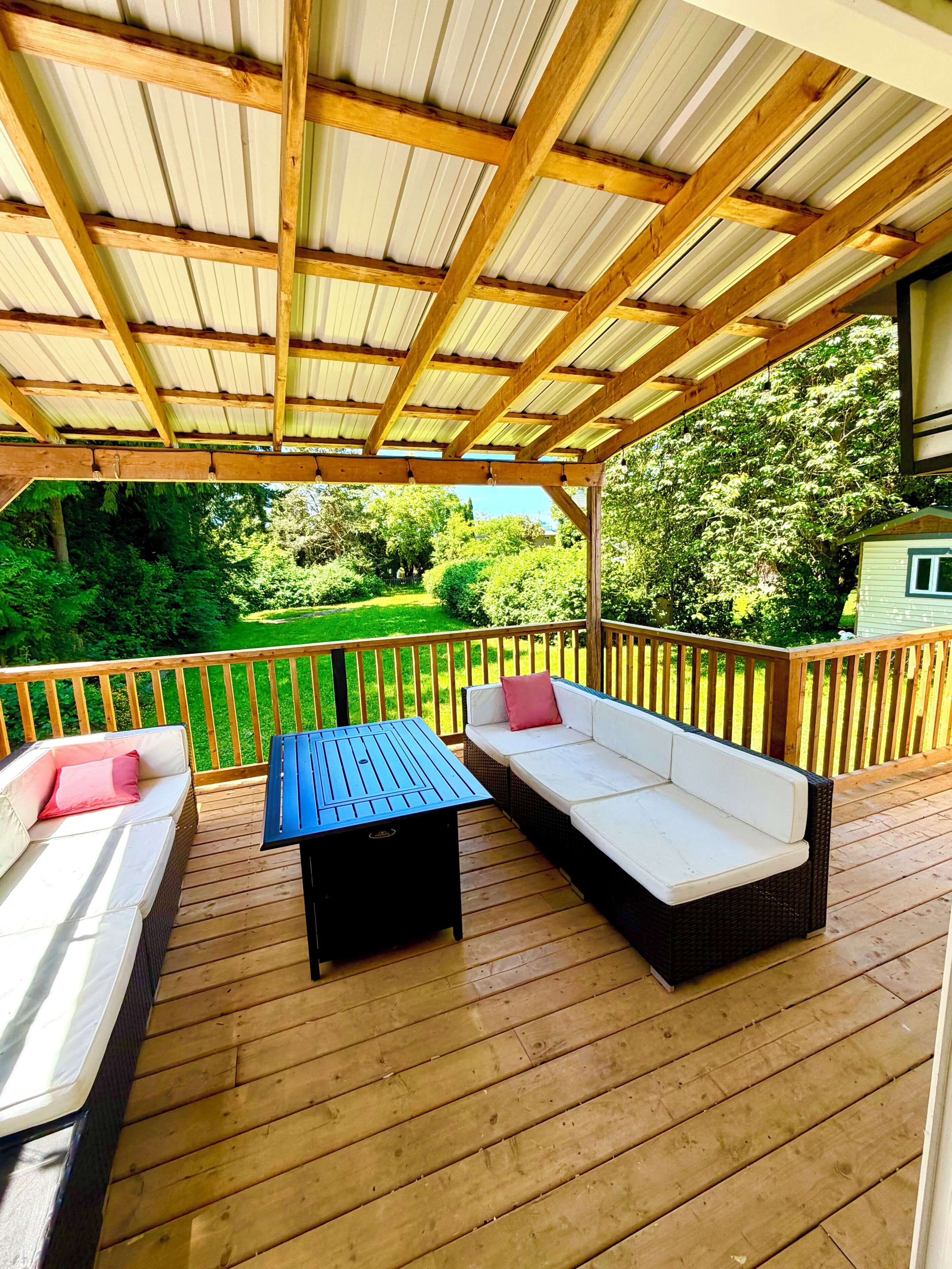 A covered deck with wooden flooring, a table, two sofas, and a view of a green landscape.