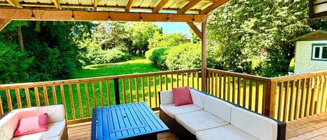 A covered deck with wooden flooring, a table, two sofas, and a view of a green landscape.