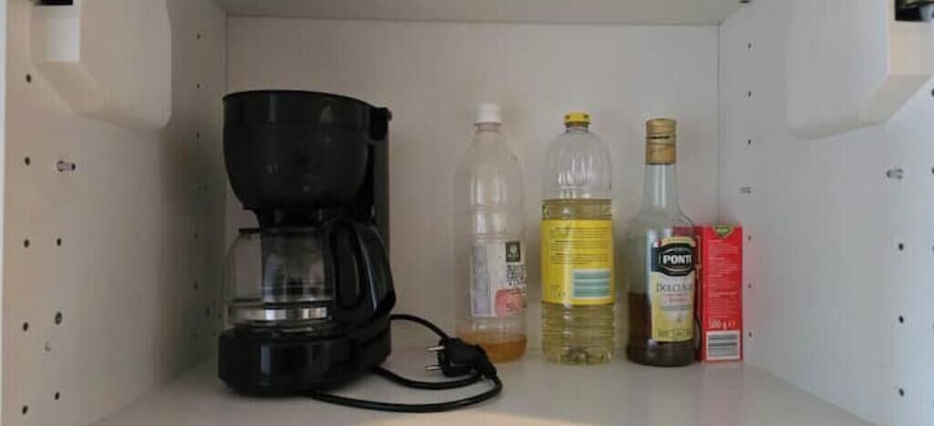A shelf with a coffee maker, a plastic bottle, a bottle of oil, and a box of sugar.