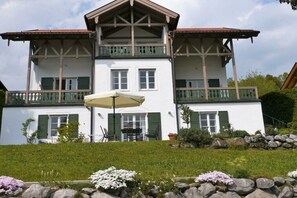A two-story building with a balcony, a dining area with outdoor furniture, and a garden with flowers and rocks. - Country House Scratches (Murnau)
