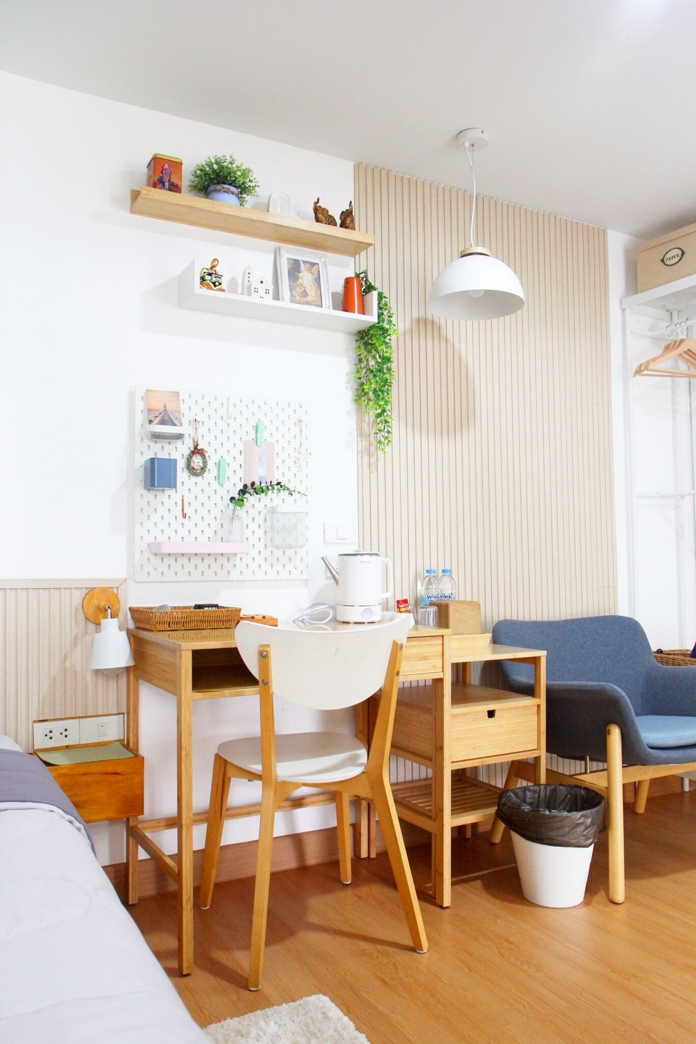 Una habitación compacta con escritorio, silla y sofá azul. Se aprecia una repisa con adornos y una planta colgante.
