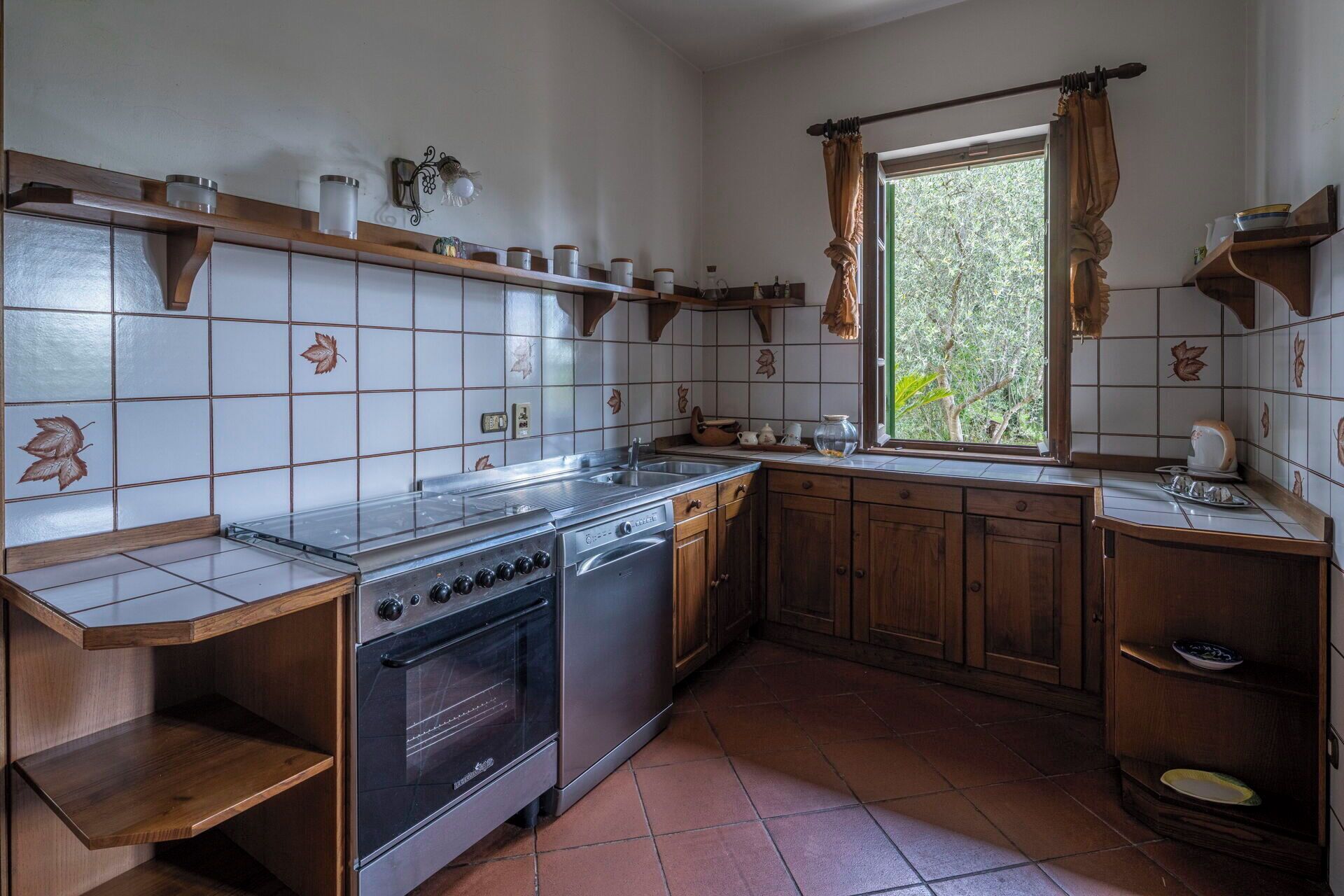 Una cocina con estufa, horno y pileta, muebles de madera y un ventanal con cortinas.