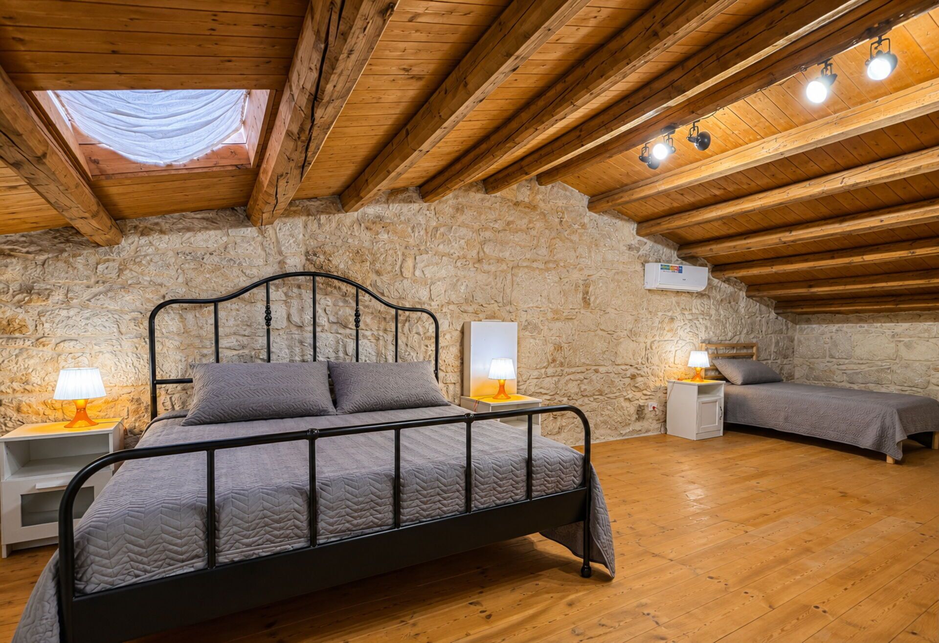 Une chambre avec un mur en pierre, des poutres en bois au plafond, un lit, des tables de chevet et des lampes.