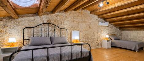 Une chambre avec un mur en pierre, des poutres en bois au plafond, un lit, des tables de chevet et des lampes.