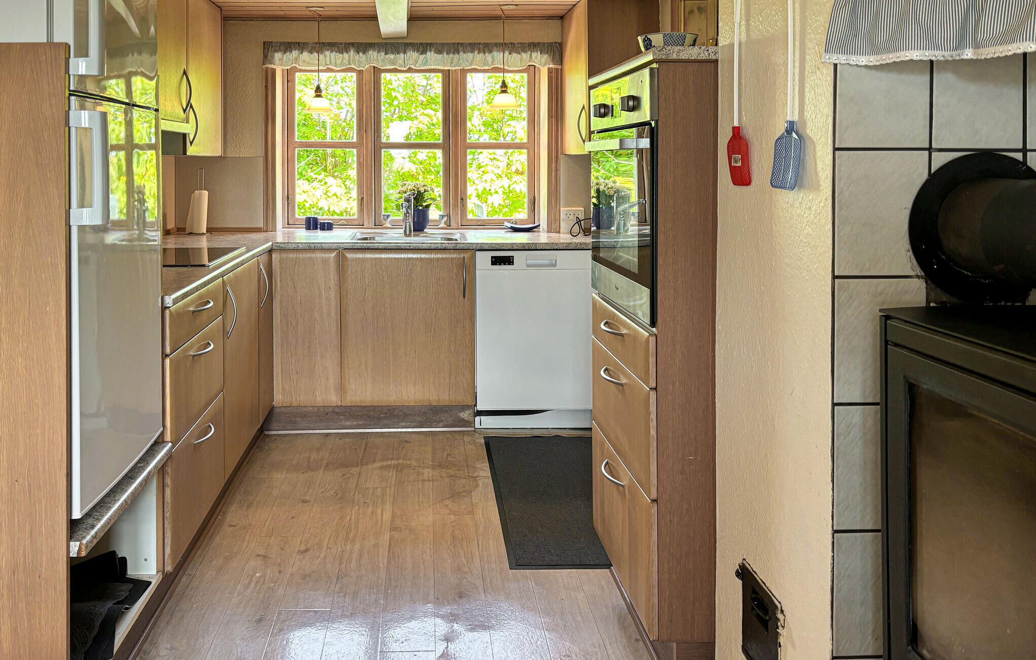 A kitchen with wooden cabinets, a white dishwasher, a window with a view of greenery, and a tiled backsplash.