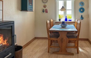 Uma sala de jantar com uma mesa de madeira, cadeiras, um fogão com fogo visível, uma janela com vista para o verde e pratos decorativos na parede.