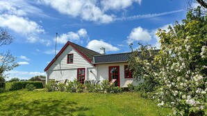 Uma casa branca com detalhes vermelhos e um telhado inclinado, rodeada por grama verde e árvores floridas.
