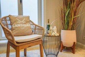 Ein Korbstuhl mit Kissen, ein kleiner runder Beistelltisch mit Flaschen und eine Champagnerflasche, sowie eine hohe Vase mit getrockneten Pflanzen.
