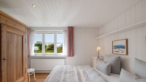 Quarto com cama, mesa de cabeceira, abajur, janela com cortinas e vista para o verde lá fora.