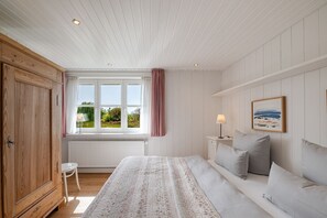 A bedroom with a bed, a nightstand, a lamp, a window with curtains, and a view of greenery outside. - Melenbarig-Hüs - Melenbarig-Hüs - Nature, sea view & relaxation on Sylt (Sylt-Ost)