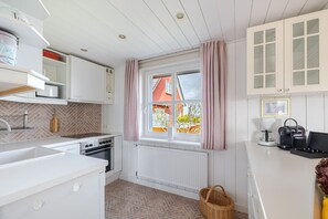 A kitchen with white cabinets, a window with curtains, a radiator, and a basket on the floor. - Melenbarig-Hüs - Melenbarig-Hüs - Nature, sea view & relaxation on Sylt (Sylt-Ost)
