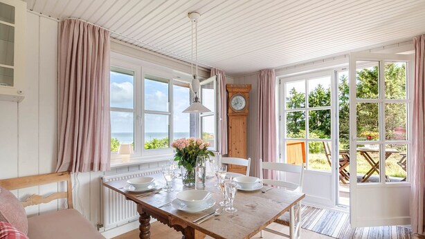Uma sala de jantar com uma mesa de madeira posta para quatro pessoas, um relógio na parede e vista para o exterior.