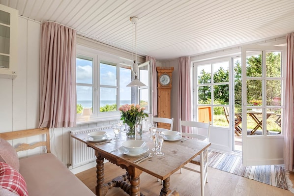 A dining room with a wooden table set for four, a clock on the wall, and a view of the outdoors. - Melenbarig-Hüs - Melenbarig-Hüs - Nature, sea view & relaxation on Sylt (Sylt-Ost)