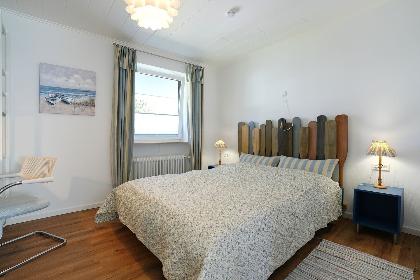 A bedroom with a bed, a chair, a nightstand, a lamp, a window with curtains, a radiator, and a painting on the wall.