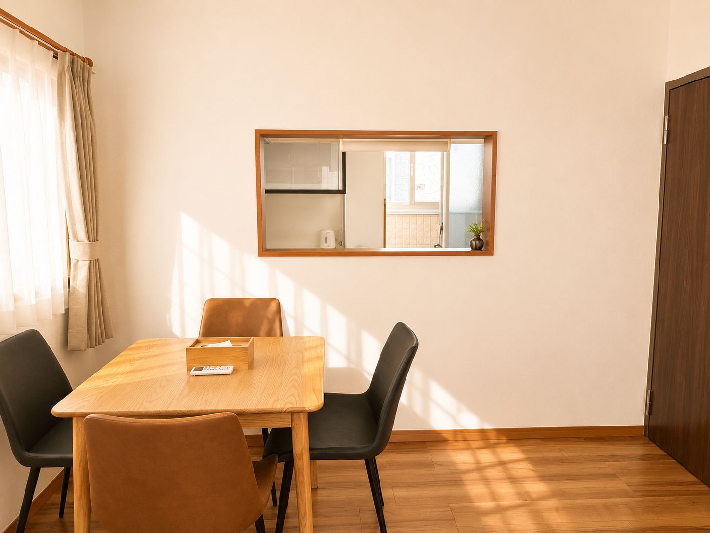 A room with a wooden dining table, two black chairs, a wooden floor, a window with a view of a kitchen, and a door.