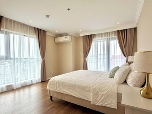 Ein Hotelzimmer mit einem großen Bett, einer Nachttischlampe und Blick auf die Stadt durch große Fenster.