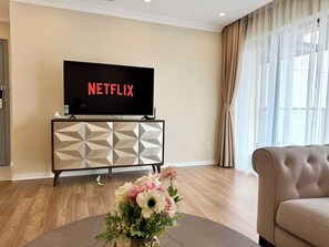 Ein Wohnzimmer mit Fernseher, der 'NETFLIX' anzeigt, einem beigefarbenen Sofa, einer dekorativen Vase mit Blumen und einem Schrank mit geometrischem Muster.