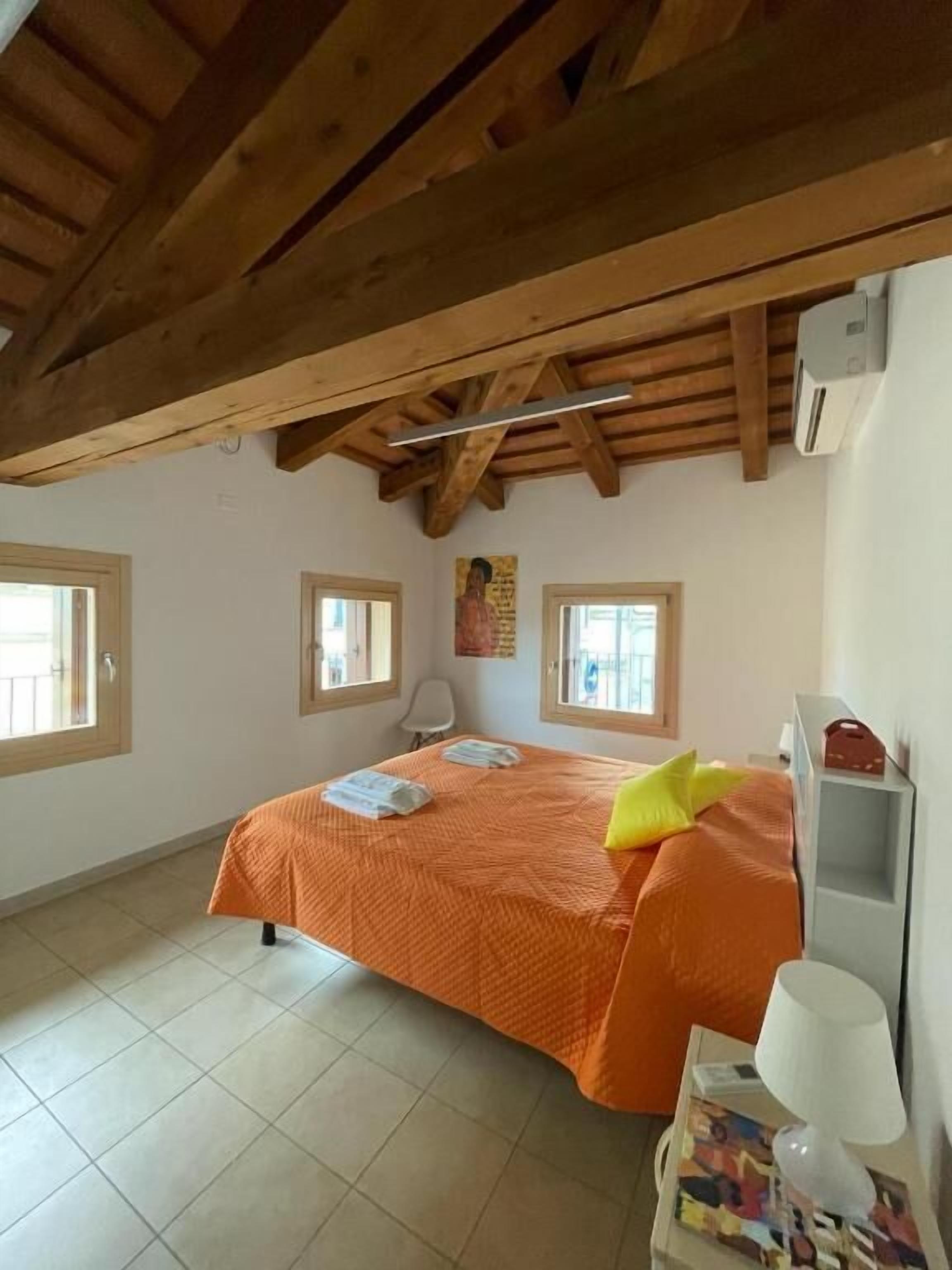 A bedroom with a wooden ceiling, a bed with an orange bedspread, two windows, and a bedside table with a lamp.