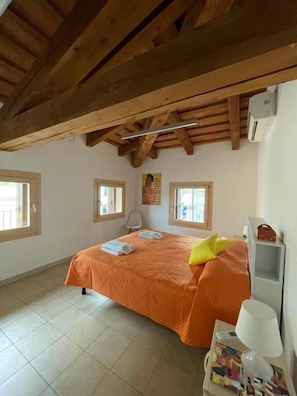 Een slaapkamer met een houten plafond, een bed met een oranje dekbedovertrek, twee ramen en een nachtkastje met een lamp.