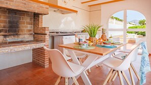 An outdoor kitchen with a brick oven, white chairs, and a dining table set with food and drinks. - Villa Dalia - Plusholidays (Calpe / Calp)