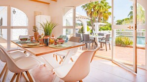 A dining area with a table set for four, surrounded by white chairs, with a view of a pool and palm trees outside. - Villa Dalia - Plusholidays (Calpe / Calp)