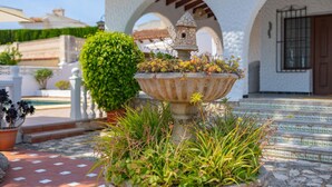 A stone fountain with a birdhouse, surrounded by plants, in front of a white building with arches. - Villa Dalia - Plusholidays (Calpe / Calp)