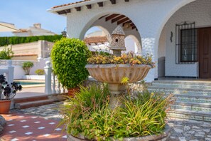 Ein Steinkonfountain mit Vogelhaus, umgeben von Pflanzen, vor einem weißen Gebäude mit Bögen.