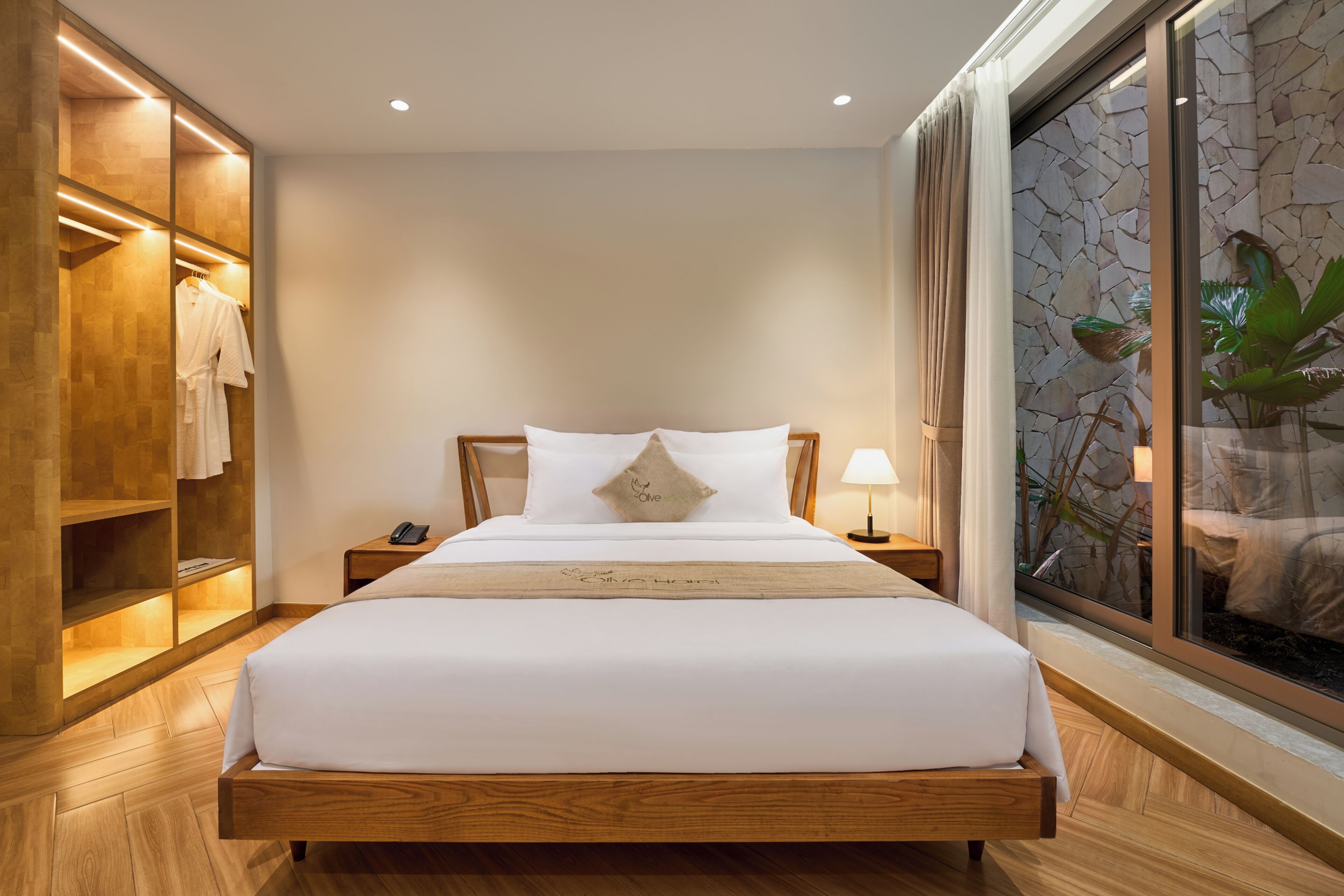 A hotel room with a wooden bed, a nightstand, a robe rack, and a large window with a stone wall view.