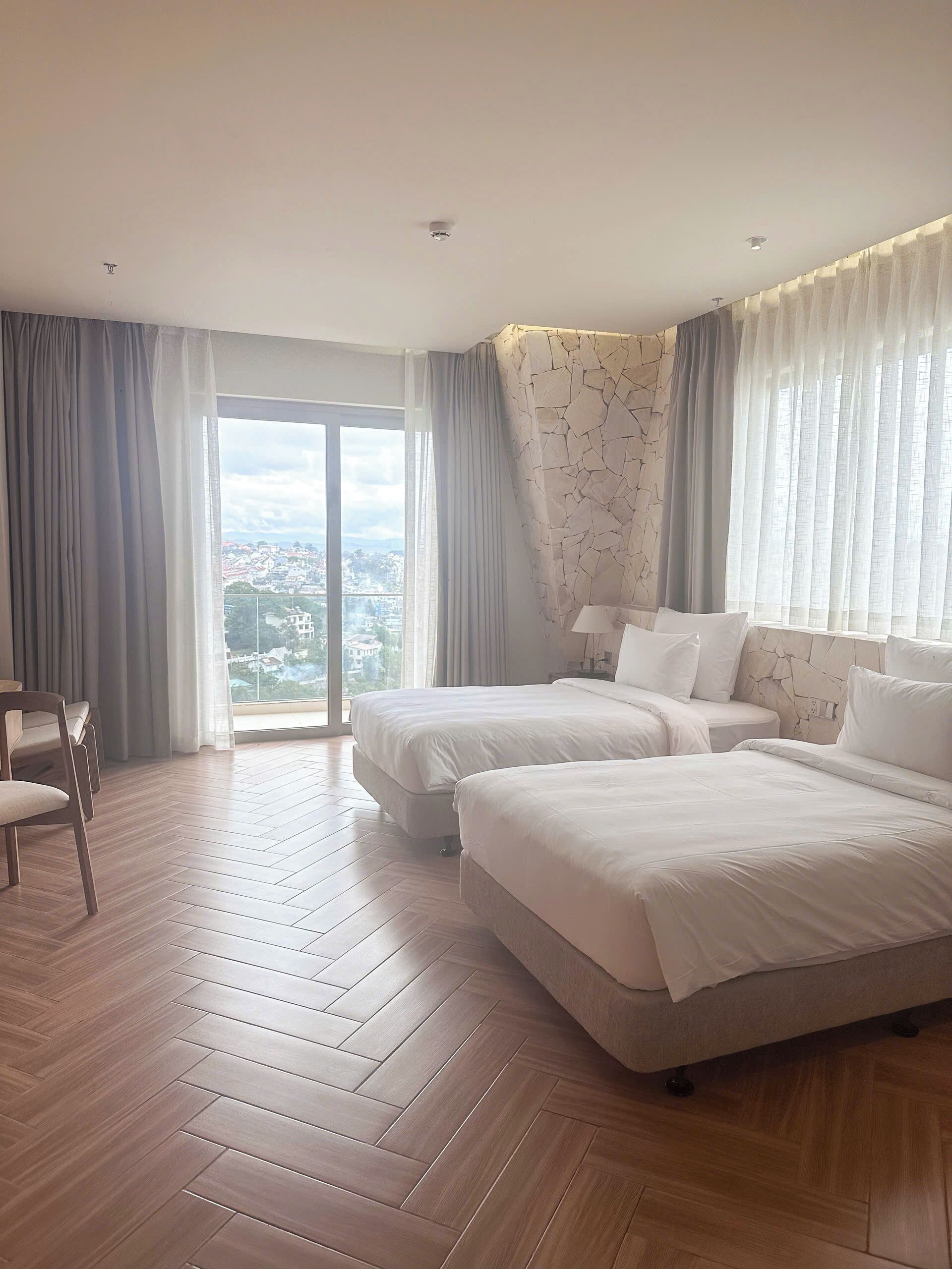 Une chambre d’hôtel avec deux lits, une chaise, une grande fenêtre avec des rideaux et une vue sur le paysage urbain.