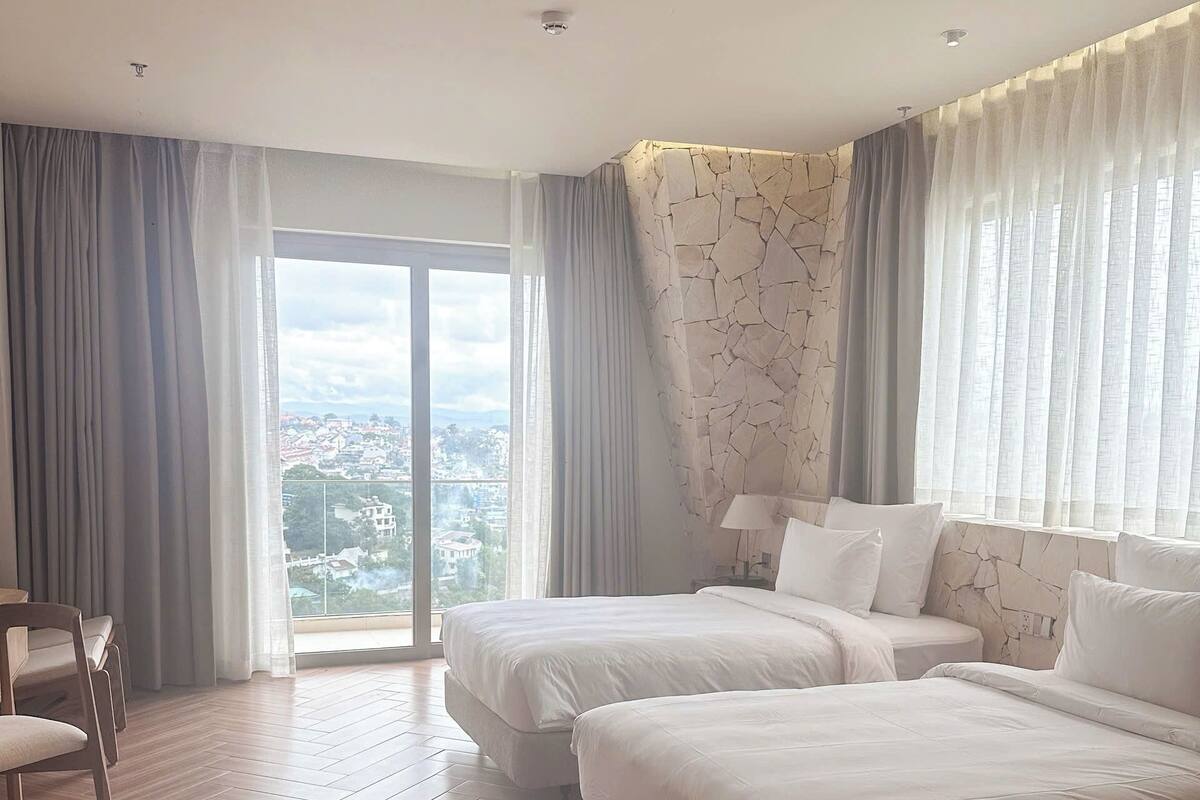 Une chambre d’hôtel avec deux lits, une chaise, une grande fenêtre avec des rideaux et une vue sur le paysage urbain.