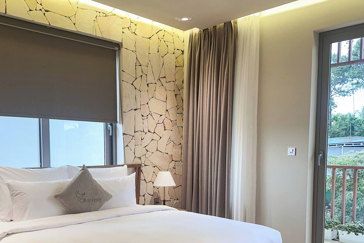 Une chambre avec un grand lit, un banc, une table de chevet, une lampe, un mur en pierre et une vue par la fenêtre.