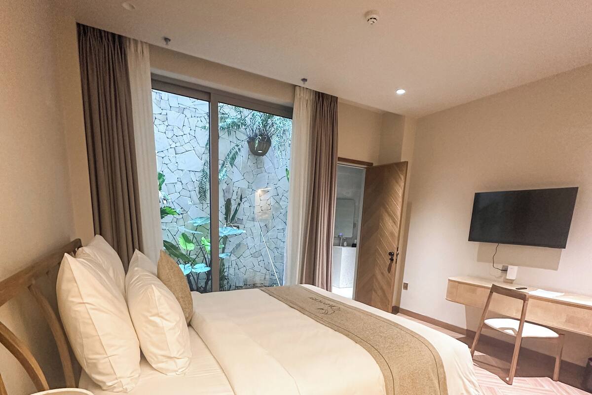 Une chambre d’hôtel équipée d’un lit, d’une télévision, d’un bureau et offrant une vue sur la verdure par la fenêtre.