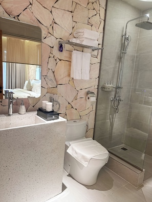 A bathroom with a stone wall, a glass shower enclosure, a toilet, a sink with toiletries, and a mirror.