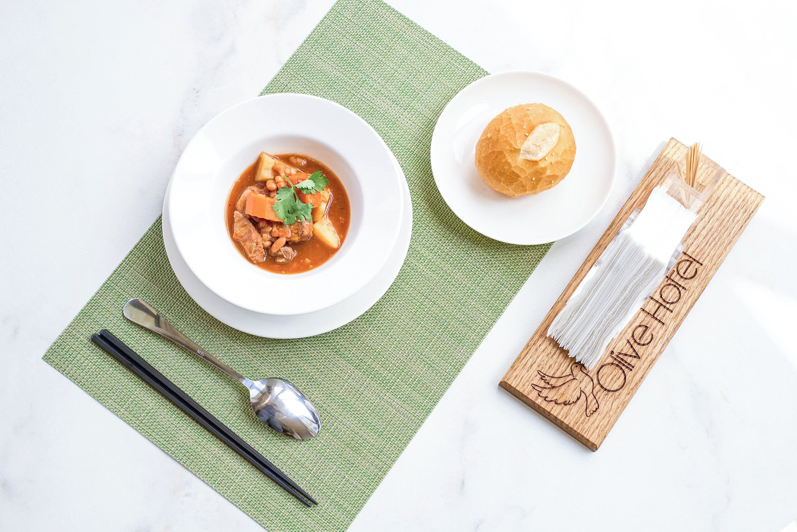 A bowl of soup, a small plate with a bun, chopsticks, a spoon, and a napkin on a green mat.