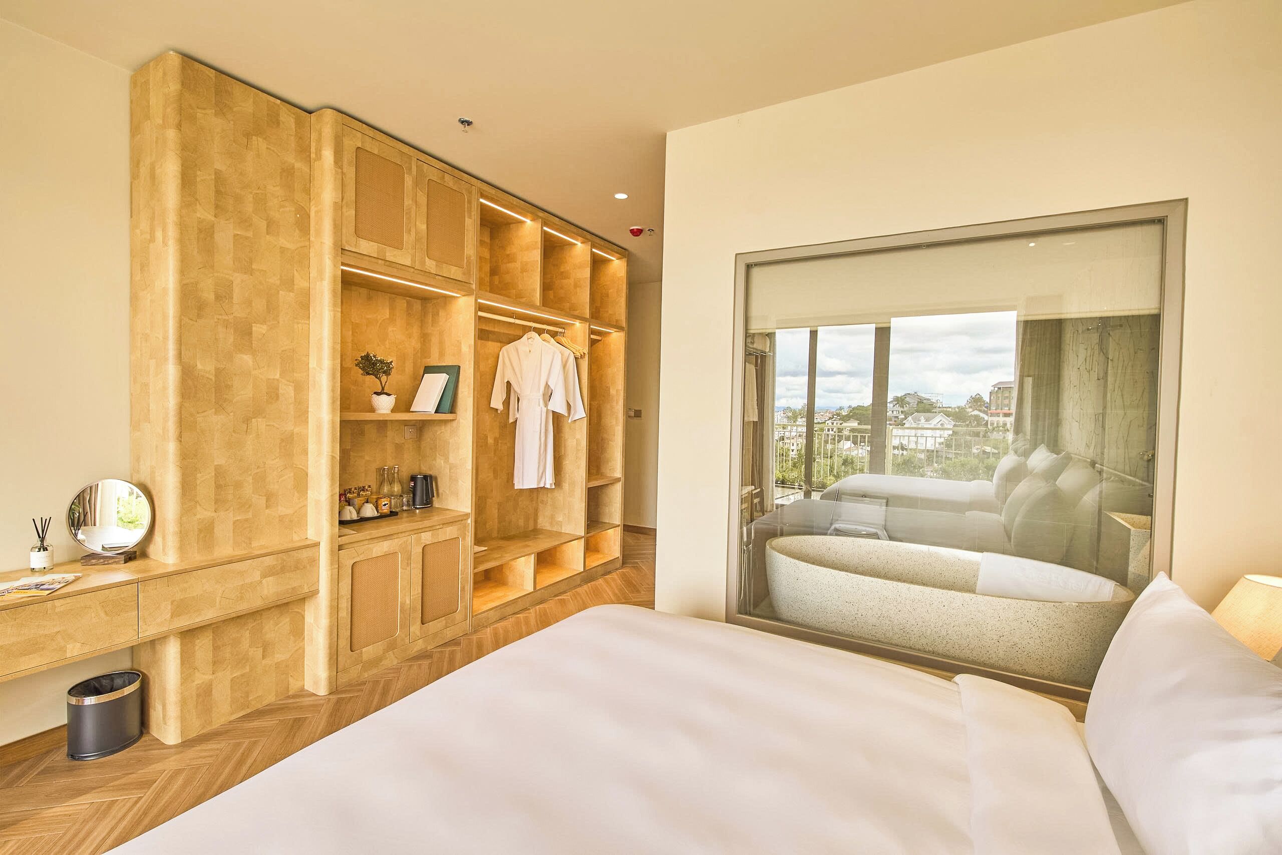 Une chambre d’hôtel avec un grand lit, une armoire avec plusieurs étagères, une petite poubelle et une fenêtre donnant sur des immeubles.