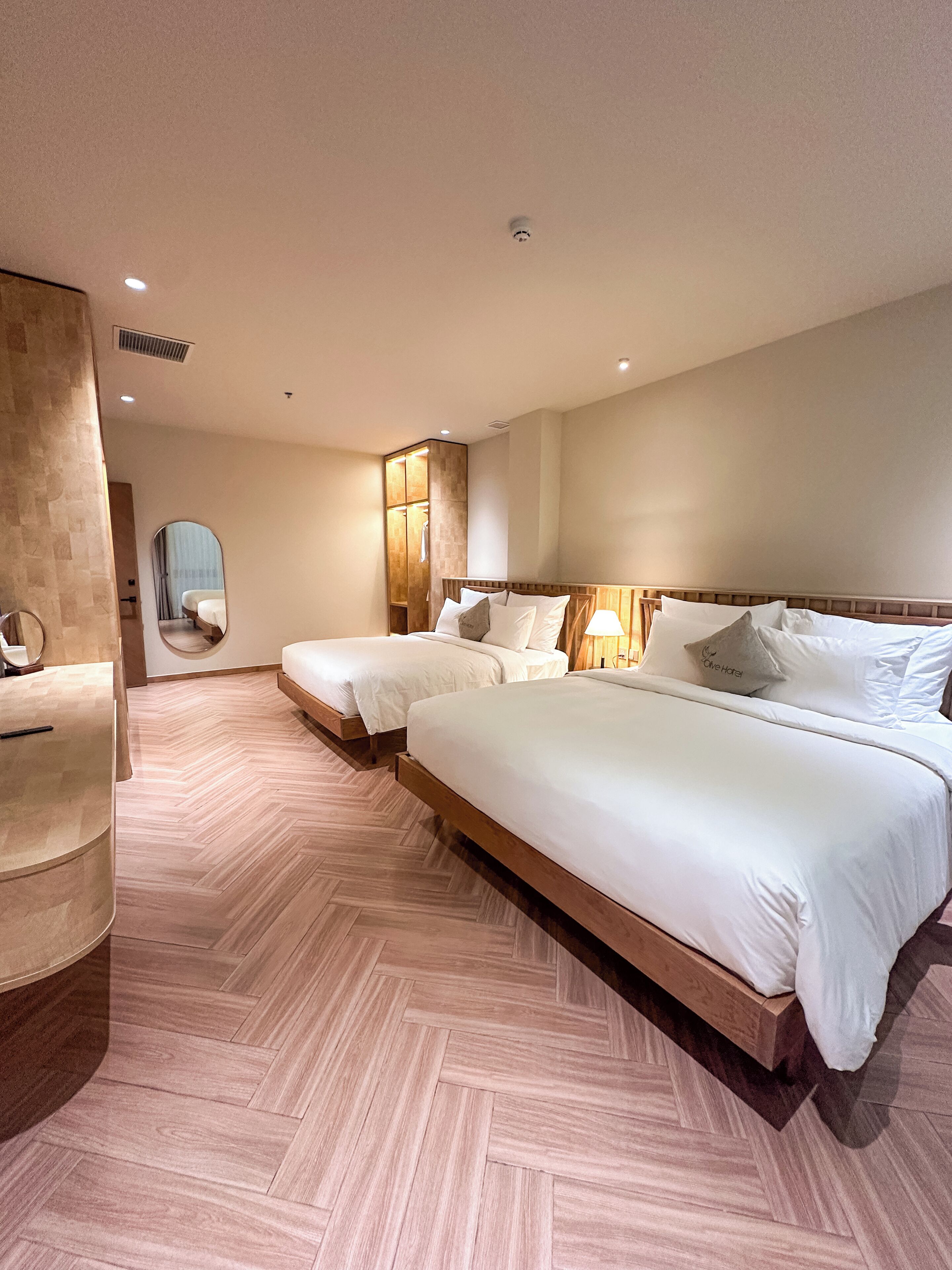 Une chambre d’hôtel avec deux lits, un miroir, une coiffeuse et un parquet à motifs en chevrons.