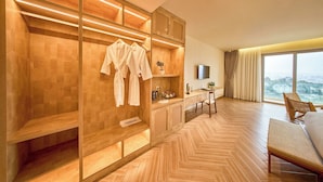 Een moderne hotelkamer met een houten visgraatvloer, een inbouw kast met ophangkappen, een klein bureau met een plant en een groot raam met uitzicht op de stad.
