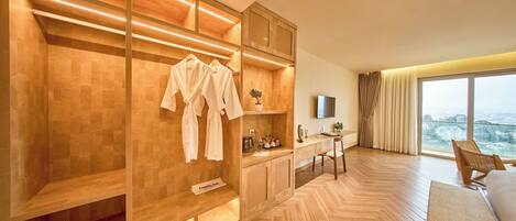 A modern hotel room with a wooden herringbone floor, a built-in wardrobe with hanging robes, a small desk with a plant, and a large window offering a city view.